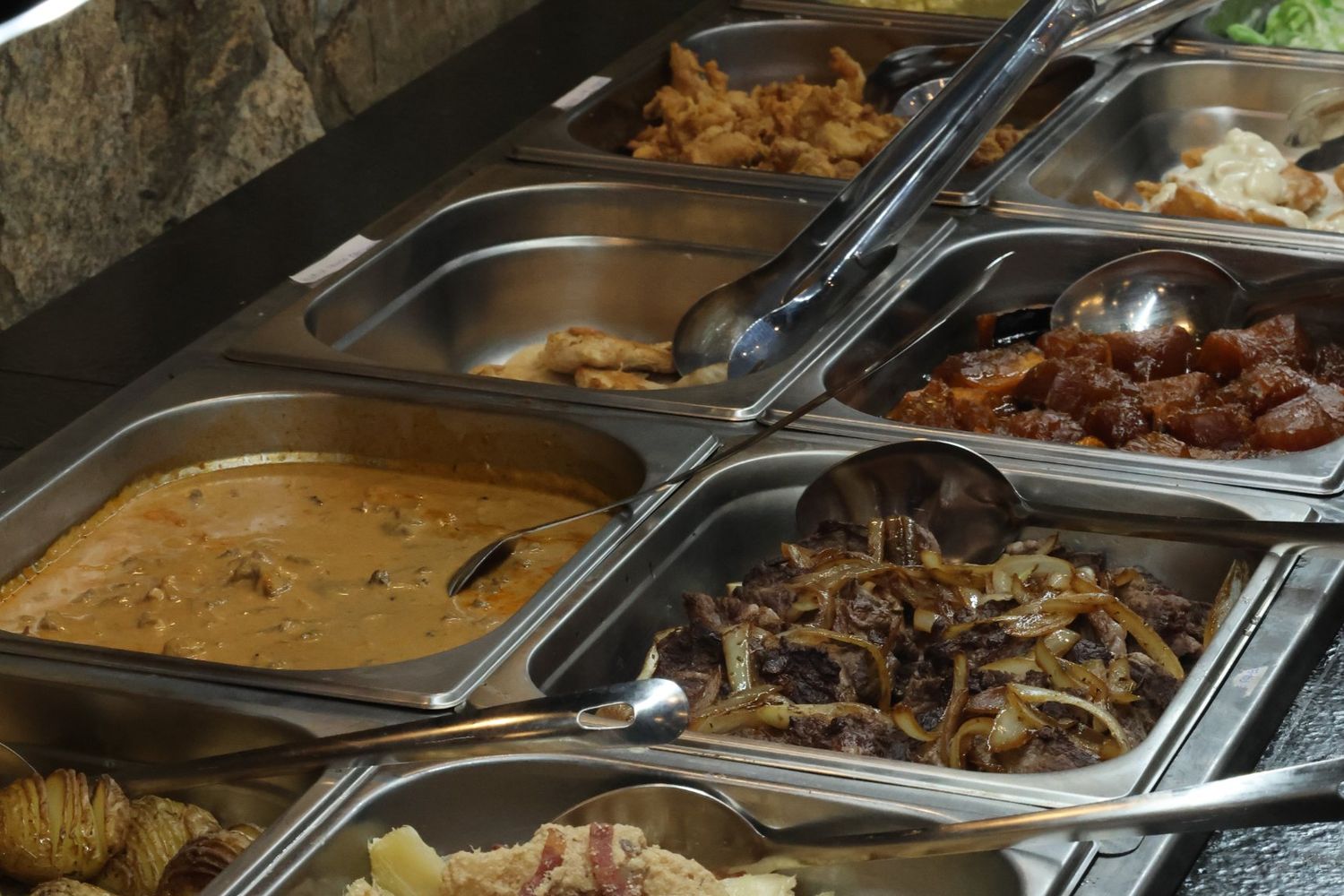 Cubas do buffet livre com pratos quentes, como frango, carne acebolada e batatas assadas