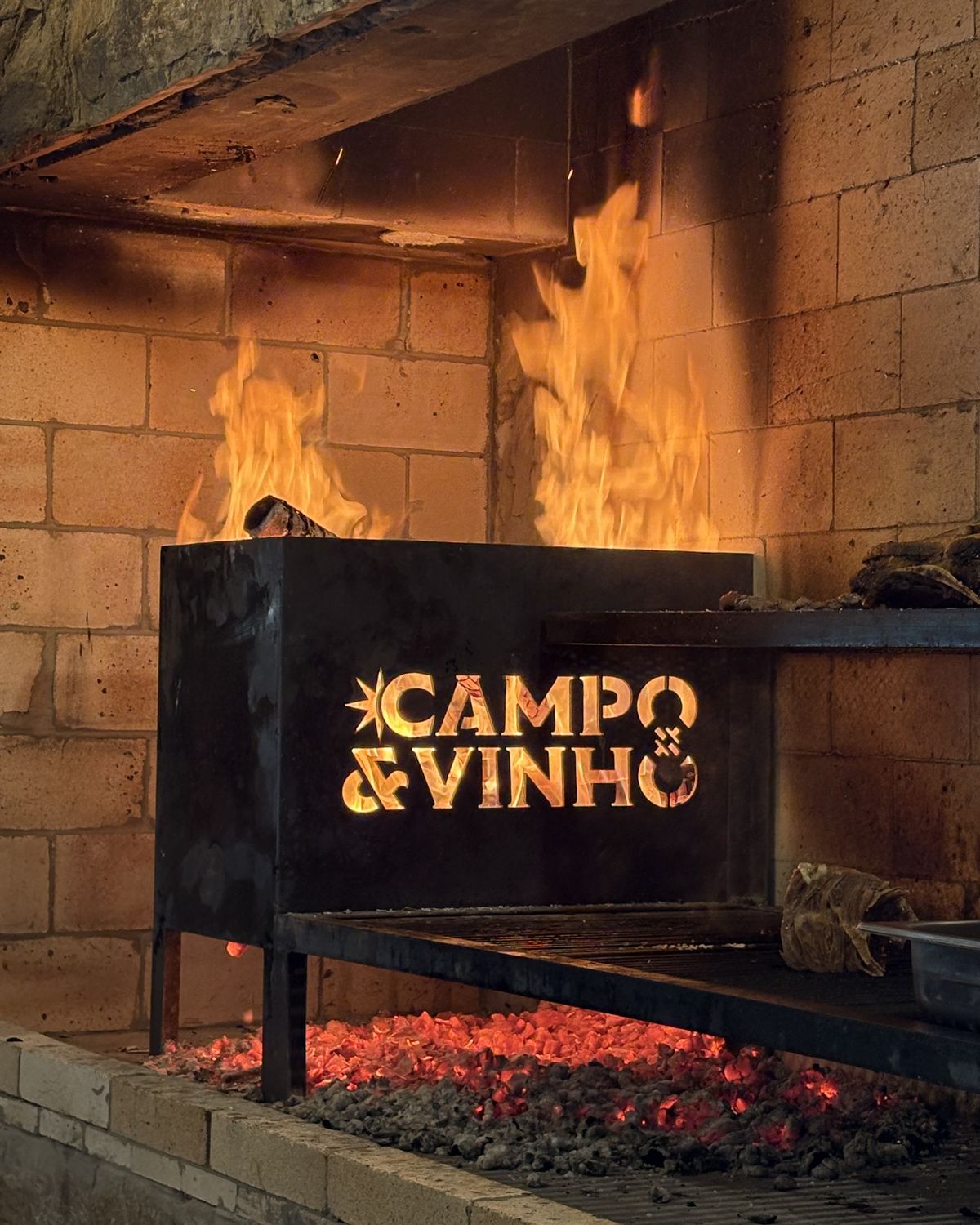 Parrilla acesa com o logo do Campo & Vinho recortado no ferro
