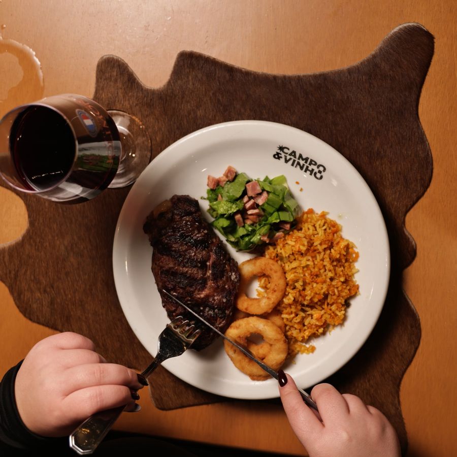 Prato com carne grelhada, salada, arroz e anéis de cebola ao lado de uma taça de vinho tinto